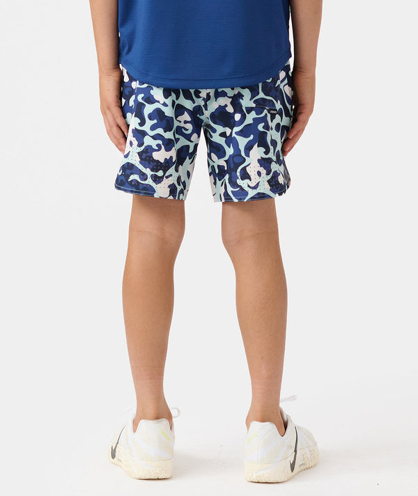 Child wearing blue t-shirt and navy, cream, and white camouflage shorts with white sneakers.