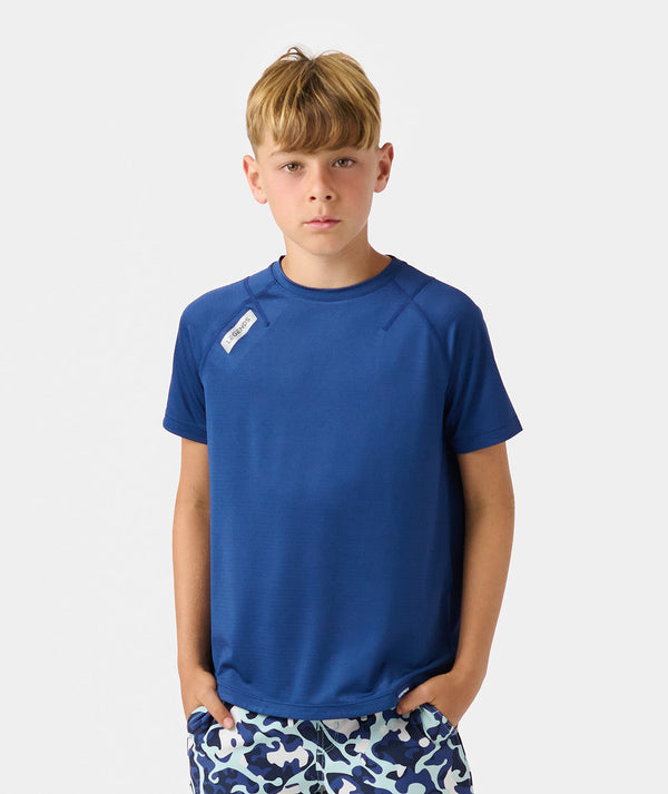 Boy wearing blue short-sleeve t-shirt with small white label on chest, paired with patterned shorts.